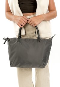 Grijze nylon draagtas met zwarte leren handvatten, gladde textuur, bovenste ritssluiting en een klein sierhangertje. Gehouden in de handen.