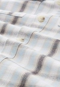 Blau-weiß kariertes Hemd mit leichter Textur; verfügt über cremefarbene Knöpfe und darke Nähte über den Stoff.