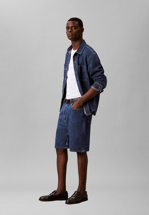 Muž stojaci s rukami v vreckách, oblečený v rifľovej bunde, bielom tričku, rifľových kraťasoch s potlačou loga a čiernych mokasínach na sivom pozadí.