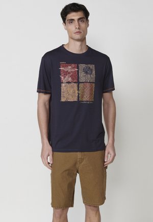 Camiseta navy con estampado gráfico que presenta cuatro patrones cuadrados distintos en rojo, beige y marrón; combinada con pantalones cortos beige.