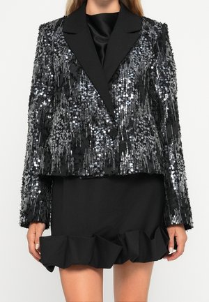 Blazer noir à sequins avec fermeture à un bouton, larges revers, porté sur une robe noire à ourlet froncé au-dessus des genoux.
