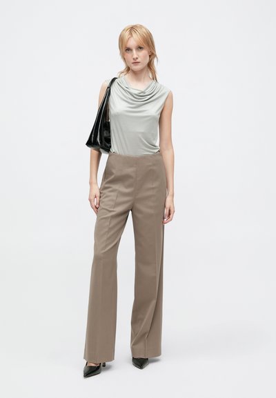Maglione grigio con scollo a cowl abbinato a pantaloni beige a vita alta con gamba larga. La modella tiene una borsa nera in vernice. Tacchi neri a punta completano il look.