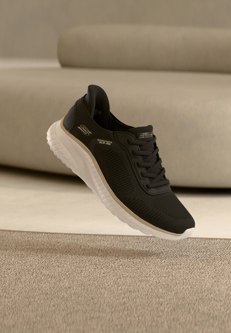 Μαύρο αθλητικό παπούτσι Skechers Slip-Ins με λευκή σόλα που αιωρείται πάνω από μπεζ χαλί μπροστά από μπεζ καναπέ.