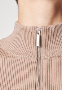 Gros plan du col d’un pull tricoté côtelé beige avec une fermeture éclair argentée partiellement fermée au niveau de l’encolure sur fond uni.