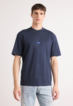 Navyblaues Baumwoll-T-Shirt mit kurzen Ärmeln und Rundhalsausschnitt, das ein kleines blaues "HUGO"-Logo auf der Brust trägt. Kombiniert mit ausgewaschenen Jeans.