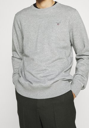 Mann trägt ein hellgraues Sweatshirt mit langen Ärmeln und einem kleinen gestickten Logo auf der Brust sowie dunkle Hose, Hände in den Taschen.