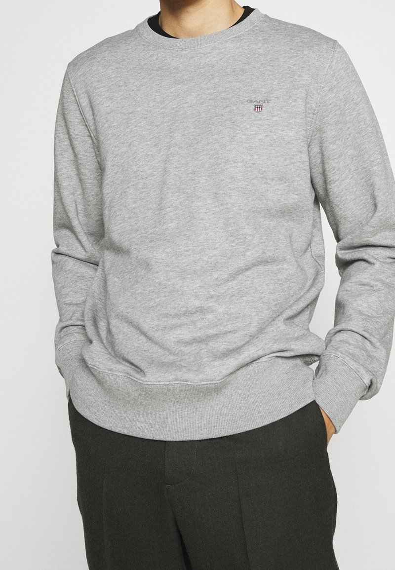 Homme portant un sweat-shirt gris clair à manches longues avec un petit logo brodé sur la poitrine et un pantalon foncé, les mains dans les poches.