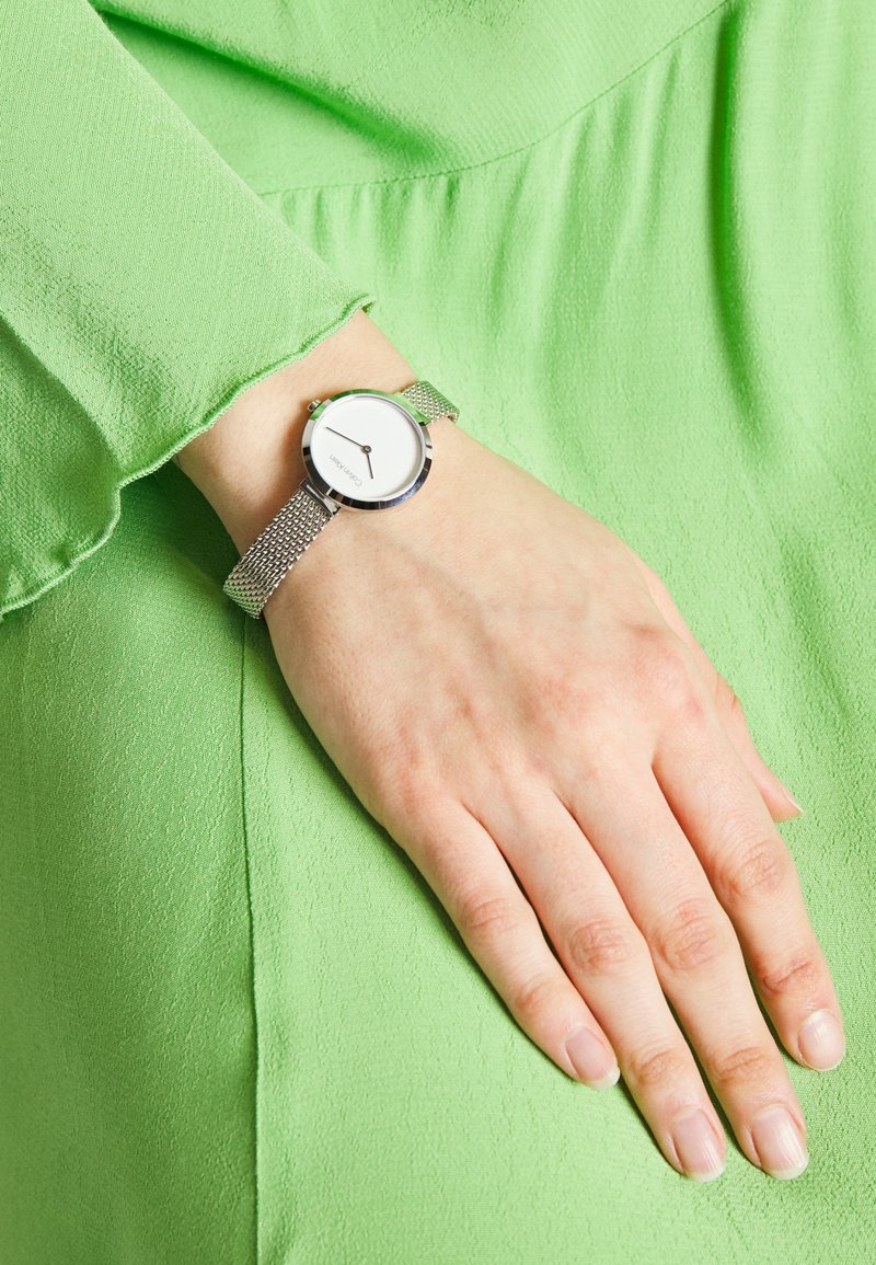 Montre avec bracelet en maille argentée et cadran rond blanc, présentant une aiguille des minutes noire simple et un accent vert sur la couronne ; portée à la main.