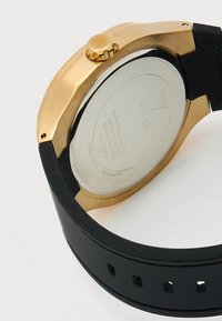 Uhrgehäuse in Goldton mit schwarzem Kautschukarmband, zeigt die Rückseite aus Edelstahl, graviert mit Marken- und Wasserdichtigkeitsangaben.