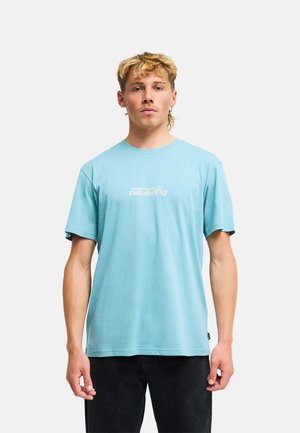 Młody mężczyzna o jasnych włosach, ubrany w jasnoniebieski t-shirt Billabong i czarne spodnie, stojący na gładkim białym tle.