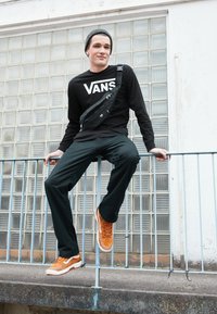 Sort langærmet skjorte med "VANS" logo, sort bæltetaske, grønne bukser, orange sneakers med hvide accenter, som sidder på en ræling.