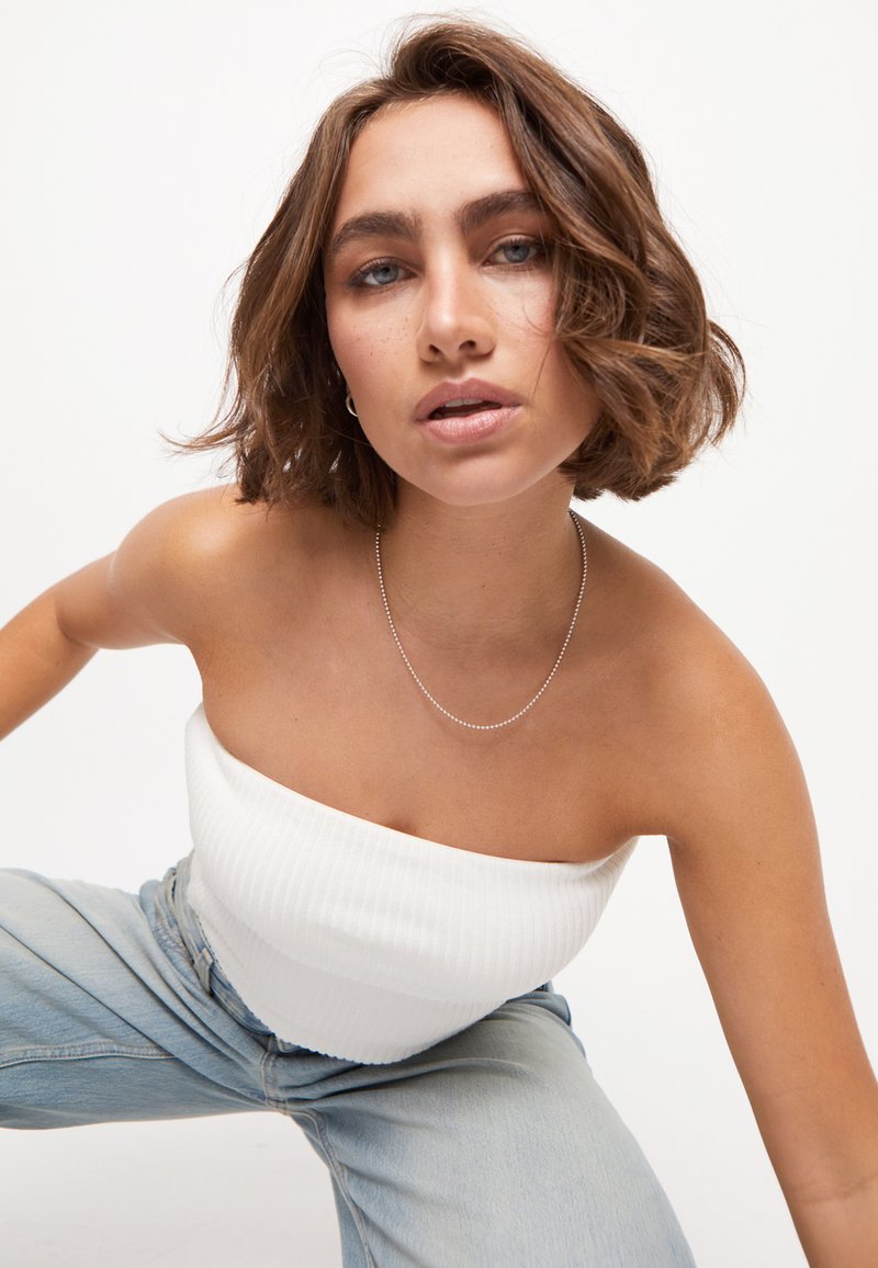 Top blanco sin tirantes con textura acanalada, collar de cadena plateada, vaqueros azul claro, pose informal, cabello con textura y ondas suaves, fondo neutro.