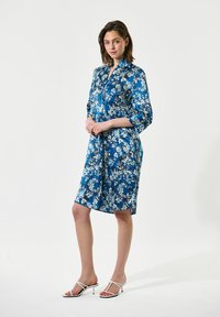 Vestito blu di media lunghezza con un motivo floreale, davanti con bottoni e maniche 3/4; abbinato a sandali bianchi con tacco. Tessuto di finitura liscia.