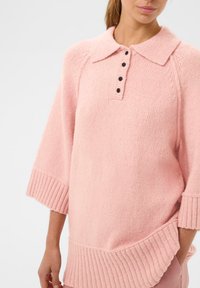 Žena v měkkém růžovém pleteném svetru s širokým límcem, čtyřmi černými knoflíky, tříčtvrtečními rukávy a žebrovanými okraji.