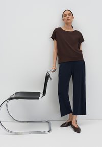 Bruine korteklaver top, marineblauwe wijde broek en donkere platte schoenen. Het model leunt op een zwarte stoel met een chroomframe.