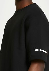 Black cotton t-shirt with short sleeves, featuring a crew neck and white text "(LÉS) ENSEMBLE" embroidered on the left sleeve.