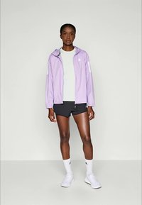 Giacca leggera viola con cappuccio, dettagli bianchi, indossata sopra una maglietta grigia e pantaloni corti neri. Modello che indossa sneakers bianche e calze corte.