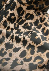 Leopardenmusterstoff mit schwarzen und tanfarbenen Mustern; verfügt über ein Logo in schwarzem Text. Glatte Textur mit einem leichten Glanz.