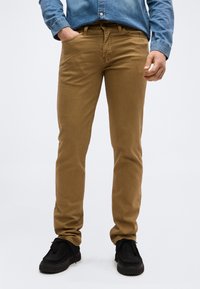 Άνδρας που φοράει καφέ στενά παντελόνια, μπλε πουκάμισο από τζιν, και μαύρα παπούτσια, στέκεται με το ένα χέρι στην τσέπη μπροστά σε απλό φόντο.