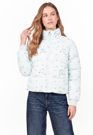 Junge Frau mit langen Haaren, die eine weiße Steppjacke mit Schneemann-Motiven und blaue Jeans trägt und mit einer Hand ihr Haar berührt.