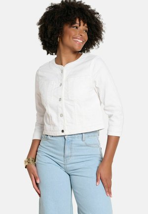 Smiling woman with curly hair wears a white buttoned jacket and light blue high-waisted jeans, accessorized with a gold bracelet.