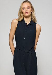 Abito senza maniche blu navy con colletto, chiusura a bottoni e cintura da annodare. Presenta un tessuto texturizzato e un piccolo logo sul petto.