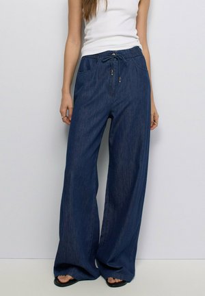 Mujer que lleva pantalones anchos de pierna ancha sueltos de color azul oscuro con cintura elástica y cordón, combinados con una camiseta sin mangas blanca y sandalias negras.