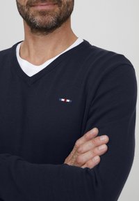 Marineblå V-hals genser laget av ribbestrikket stoff, lagt over en hvit T-skjorte, med en liten brodert logo i rødt, hvitt og blått.