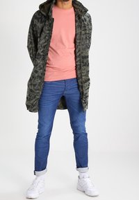 Kamouflage-mönstrad kapuschongjacka över en rosa skjorta, blå jeans och vita sneakers. Jackan har en avslappnad passform och når till mitten av låren.