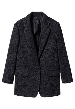 Personne portant un blazer en chevrons foncé, une chemise blanche, un foulard noir à motifs au cou, un pantalon noir, main dans la poche, avec des bagues en or à la main droite.