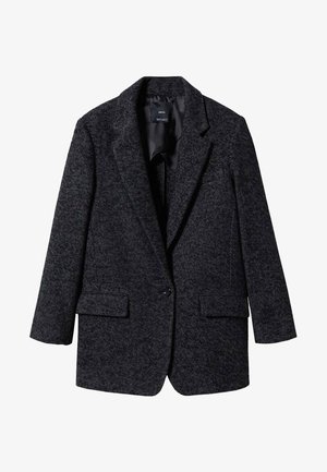 Persona che indossa un blazer scuro a spina di pesce, camicia bianca, sciarpa nera a motivi al collo, pantaloni neri, mano in tasca, con anelli d'oro sulla mano destra.