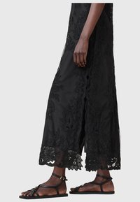 Bas du corps d'une personne portant une jupe midi en dentelle florale noire et des sandales plates noires à brides sur un fond uni.