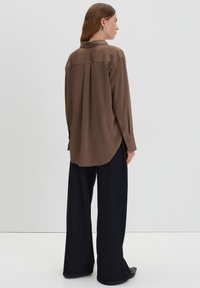 Bruine blouse met lange mouwen en een gladde textuur, voorzien van een kraag en een plooitje aan de achterkant. Draagt bij bredere zwarte broek en platte zwarte schoenen.