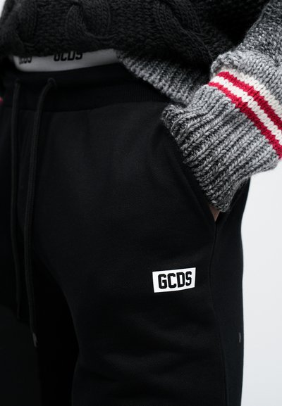 Schwarze GCDS-Sweatpants mit einem weißen Logopatch, elastischem Bund und Seitentaschen, kombiniert mit einem grauen Strickpullover mit roten Streifen.