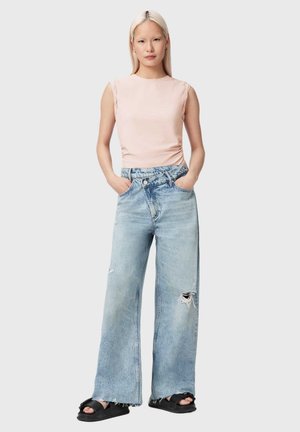 Nainen seisoo käsineen taskuissa, pukeutunut hihattomaan vaaleanpunaiseen toppiin, leveälahkeisiin, revittyihin vaaleansinisiin farkkuihin ja mustiin sandaaleihin yksiväristä taustaa vasten.