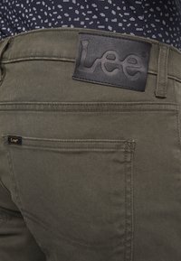 Primer plano de pantalones de la marca Lee color verde oliva, mostrando el bolsillo trasero y una etiqueta de cuero negro con el logo de Lee sobre la cintura.