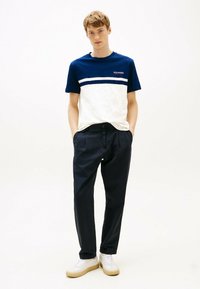 Camiseta bloqueada en color azul marino y blanco con cuello redondo, que presenta un logo. Combinada con pantalones oscuros de pinzas azul marino y zapatillas blancas.