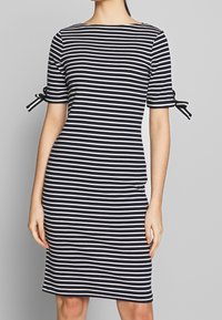 Vestido a rayas blancas y negras con escote redondo, mangas cortas y detalle de lazo en los puños. Diseño ceñido al cuerpo, longitud hasta la rodilla.