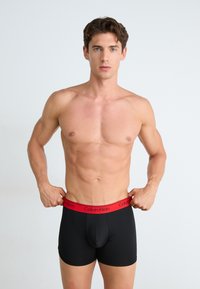 Boxeri negri din material elastic, cu un brâu roșu îndrăzneț pe care este inscriptionat logo-ul "Calvin Klein". Model ajustând brâul.
