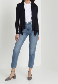 Eine Frau in einem weißen Hemd, einem schwarzen offenen Cardigan, hochtaillierten blauen Jeans und grauen Schuhen mit Knöchelriemen steht vor einem einfarbigen Hintergrund.