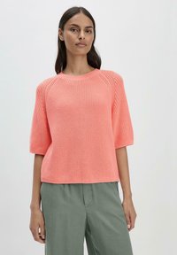 Maglione a maniche corte, lavorato a maglia, in un vivace corallo, con un motivo testurizzato e un collo rotondo. Abbinato a pantaloni leggeri e morbidi di colore verde chiaro.
