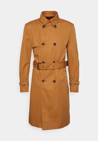 Cappotto trench marrone realizzato in tessuto liscio, con una chiusura doppiopetto, cintura e polsini con bottoni. Collo classico con cuciture in evidenza.