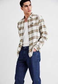 Noor mees kannab valget ja kollast ruudulist särki valge topi peal tumedate siniste teksadega, käed taskutes, vaadates kõrvale.