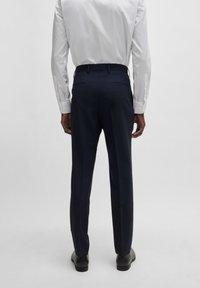 Marineblaues Anzug-Hosen mit tailliertem Schnitt, glattem Stoff und leichtem Glanz. Verfügt über Gürtelschlaufen und einen Knopfverschluss. Getragen mit einem weißen Hemd.