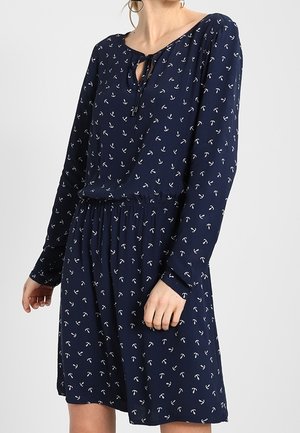 Robe bleu marine à manches longues avec motif d'ancres blanches, taille élastique et col avec nœud devant, portée par une personne debout.