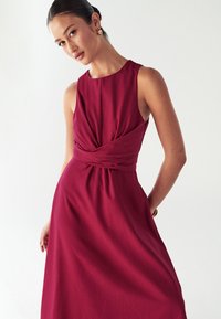 Abito senza maniche di colore bordeaux con un design intrecciato in vita, gonna fluida e texture del tessuto liscia. I dettagli discreti sulla schiena aggiungono interesse.