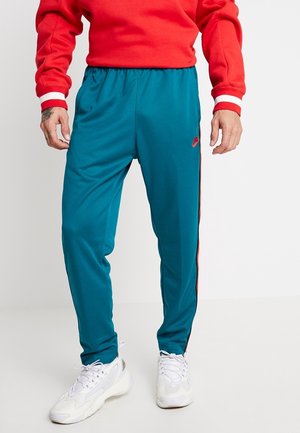 Homme portant un pantalon de survêtement bleu sarcelle avec un logo rouge, des baskets blanches, et un sweat-shirt rouge avec des poignets blancs, debout devant un fond blanc uni.