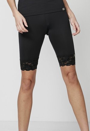 Kvinde iført sorte knælange leggings med blomsterblondekant forneden, stående med let spredte ben mod en ensfarvet baggrund.
