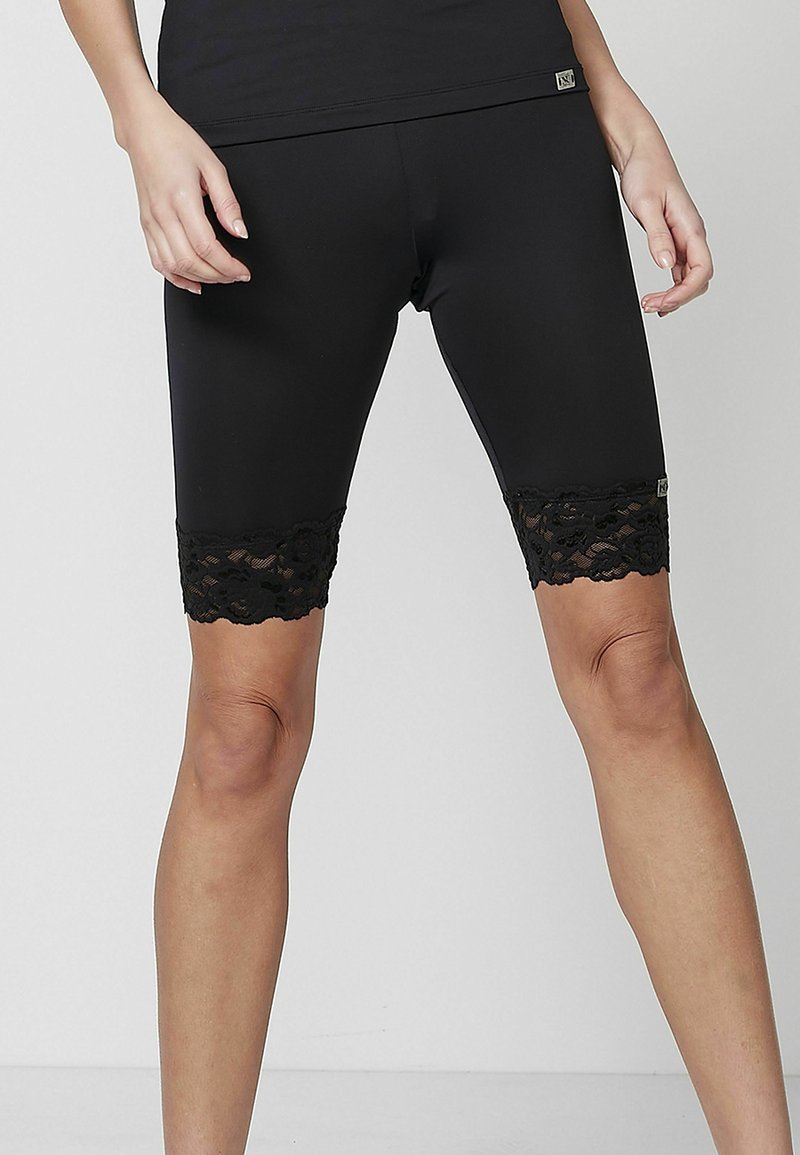 Femme portant des leggings noirs mi-genoux avec une bordure en dentelle florale à l'ourlet, debout jambes légèrement écartées sur un fond uni.