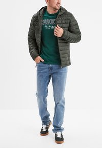 Chaqueta acolchada verde oliva con capucha, llevada sobre una camiseta verde oscura. Combinada con jeans de un azul claro y zapatillas negras con cordones blancos.
